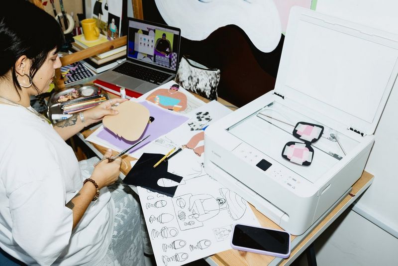 Collagekunstenaar Beth Fraser knipt een stuk karton uit. Er liggen verschillende papieren voorwerpen voor haar, er staat een laptop op de achtergrond en een multifunctionele Canon PIXMA-printer naast haar.