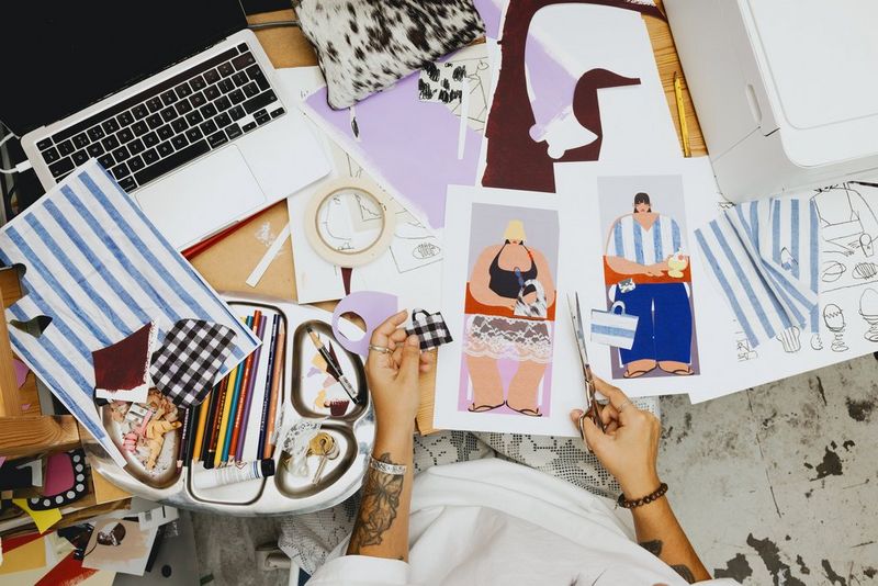 Een bovenaanzicht van collagekunstenaar Beth Fraser aan het werk. Ze houdt een schaar in de ene hand en een uitgeknipte handtas met patroon in de andere, met verschillende vellen papier, lopende projecten en tekenspullen op tafel.