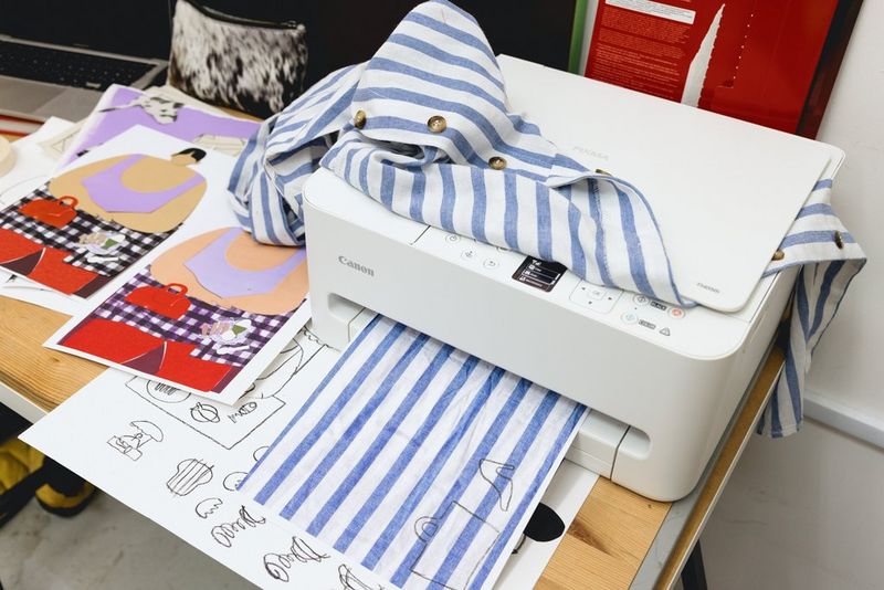 Een gestreept shirt ligt op de glasplaat van een multifunctionele Canon PIXMA-printer terwijl een print van het patroon eronder verschijnt. Op de tafel liggen werken van collagekunstenaar Beth Fraser.