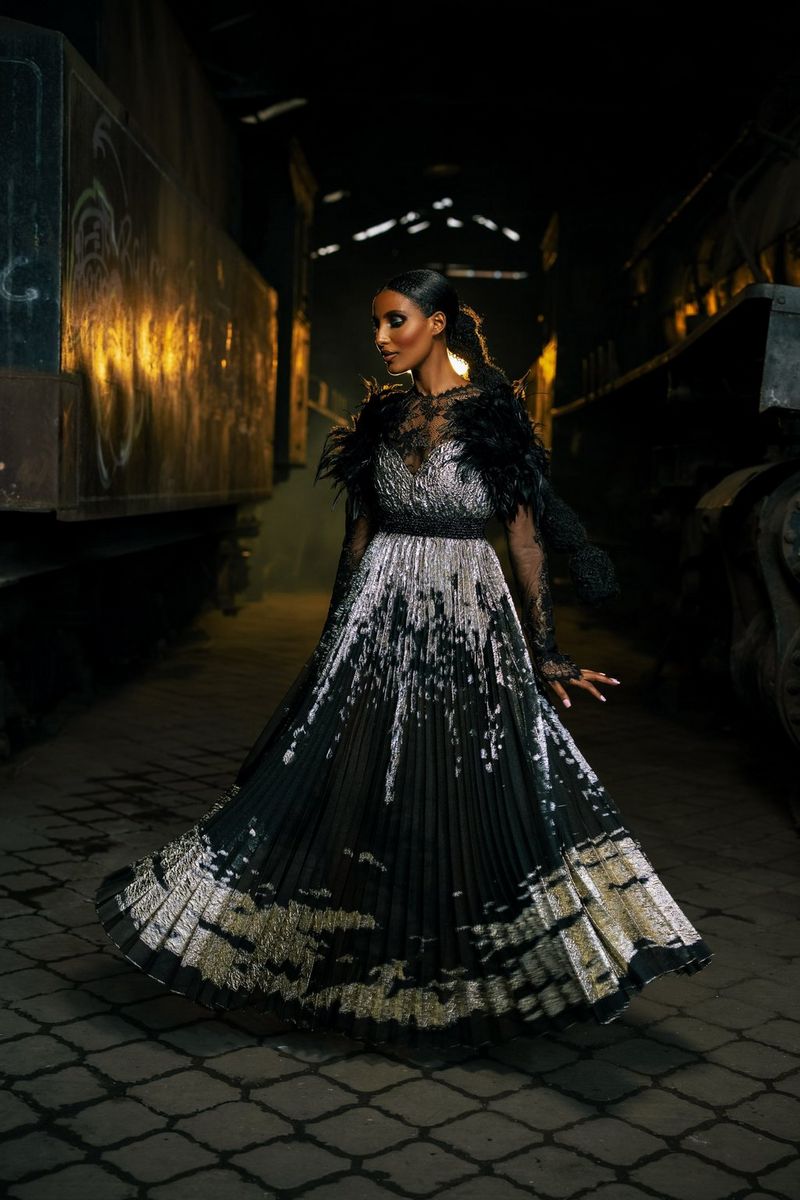 Un model într-o rochie neagră lungă, cu corsaj argintiu texturat şi motive argintii pe şolduri şi în partea de jos care flutură, într-o fotografie de modă realizată într-un hangar vechi de Emmanuel Oyeleke, cu un aparat foto Canon EOS R5 cu obiectiv Canon RF 50mm F1.2L USM.