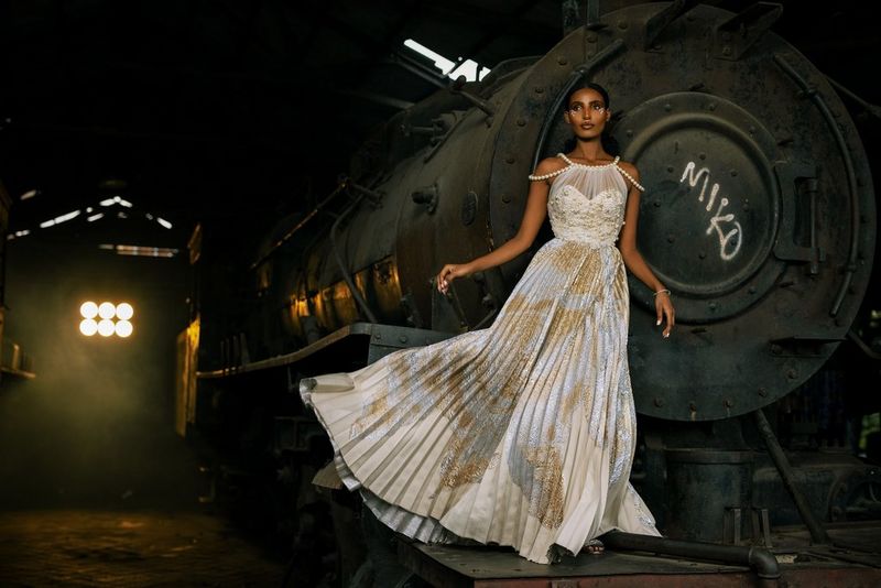 Un model purtând o rochie albă lungă, cu motive aurii şi curele din perle, stă în picioare pe un tren cu abur ruginit într-un hangar vechi, cu partea de jos a rochiei fluturând. Fotografie de modă realizată de Emmanuel Oyeleke cu un aparat foto Canon EOS R5 cu obiectiv Canon RF 50mm F1.2L USM.