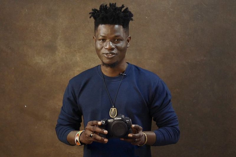 Fotograful de modă Emmanuel Oyeleke aflat în faţa unui fundal de studio în nuanţe arămii, ţinând în mână un aparat foto Canon EOS R5.