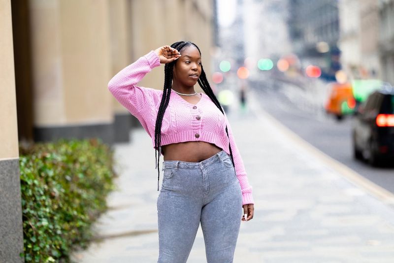 אישה עם צמות ארוכות מצטלמת על מדרכה לבושה חולצת סריג חתוכה ורודה וג'ינס אפור בגזרה גבוהה.