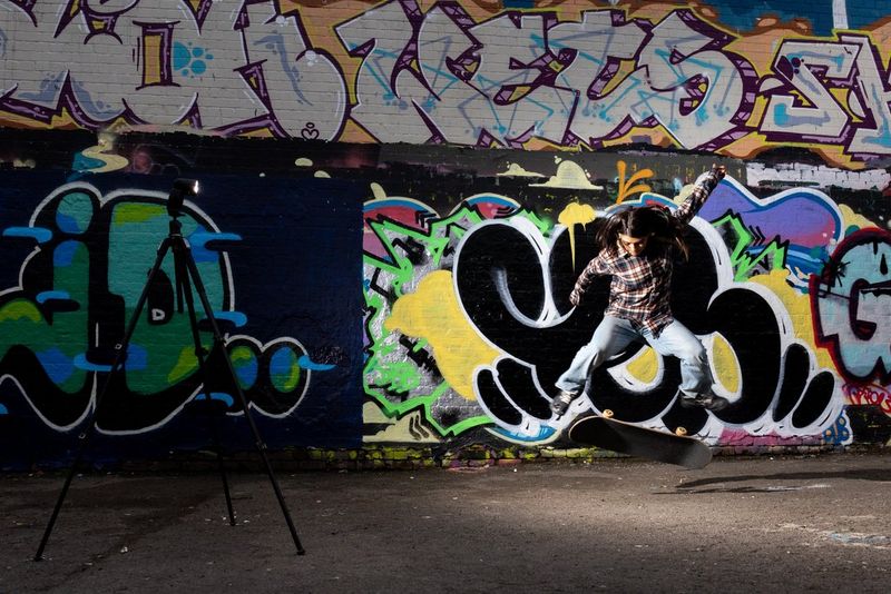 Una skateboarder che esegue un trick davanti a un muro ricoperto di graffiti, illuminata da un flash Speedlite su un treppiede.