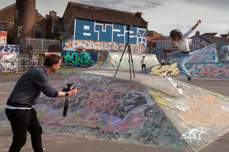 Un uomo che fotografa una donna che esegue un trick sullo skateboard, la sua tavola appare capovolta nello scatto mentre salta.
