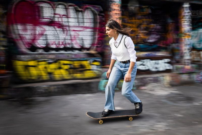 Snímek ženy na skateboardu, na jehož pozadí je rozostřená cihlová stěna pomalovaná graffiti, zatímco žena je zaostřená. Pořízeno fotoaparátem Canon EOS RP s objektivem Canon RF 24–105 mm F4–7,1 IS STM a bleskem Canon Speedlite 600EX II-RT při nastavení 26 mm, 1/30 s, f/16 a ISO 100.