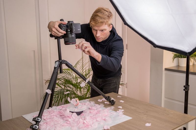 Een man zet een camera op een statief en richt hem omlaag. Hij fotografeert een roze horloge op een ronde, witte plaat waar roze rozenblaadjes omheen dwarrelen.