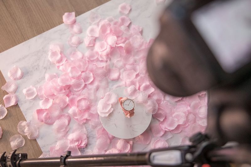 Een camera op een statief is recht omlaag gericht om een flatlay-foto van een roze horloge te maken. Het horloge ligt op een ronde, witte plaat, met roze rozenblaadjes die eromheen omlaag dwarrelen.