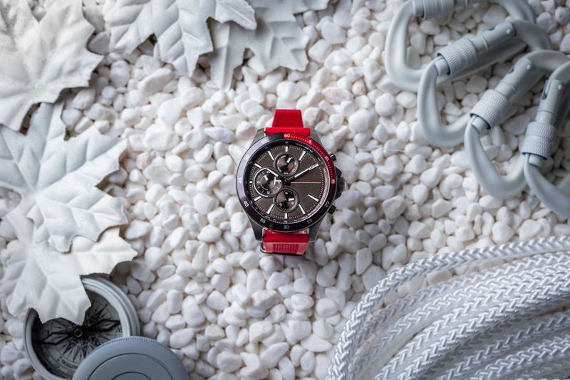 Een flatlay-foto van een rood-zwart horloge op kleine, witte stenen, met witte blaadjes en andere witte props eromheen.