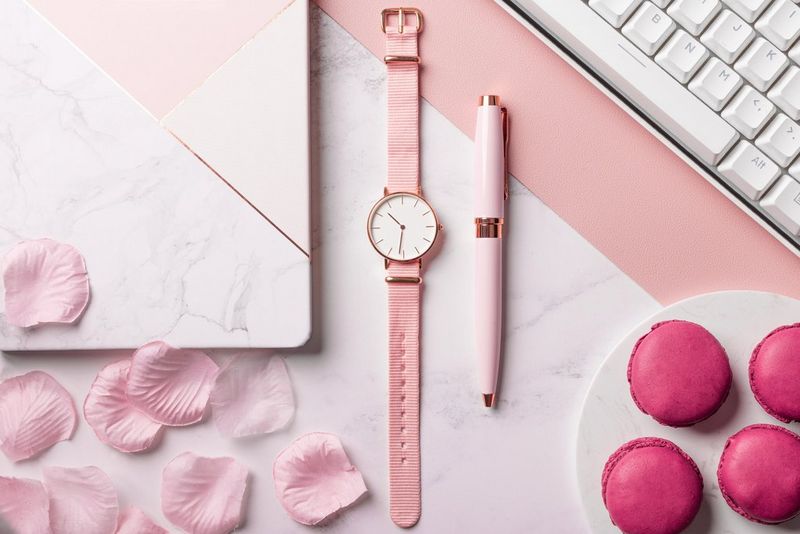 Een flatlay-foto van een roze horloge met een toetsenbord, notitieboekje, pen, bloemblaadjes en macarons die er zorgvuldig omheen zijn geplaatst, allemaal roze en wit van kleur.