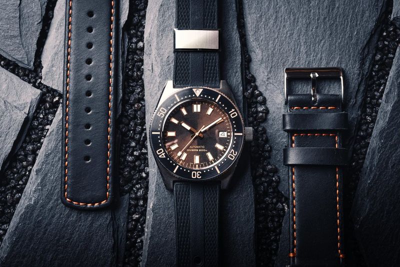 Een close-up van een flatlay-foto van een zwart horloge met zwarte bandjes aan beide kanten, op een achtergrond van gebarsten leisteen.