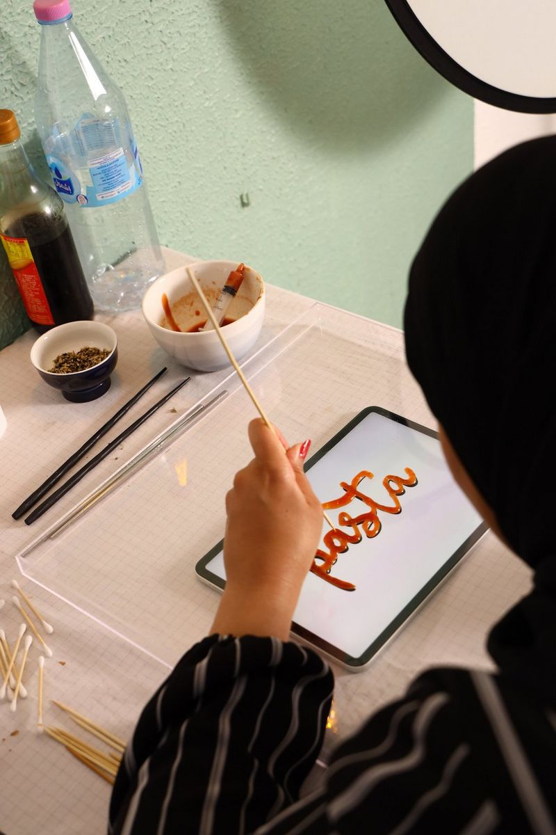 Een foto van over de schouder van foodfotograaf Yasmin Albatoul die het woord pasta tekent met een houten stokje.