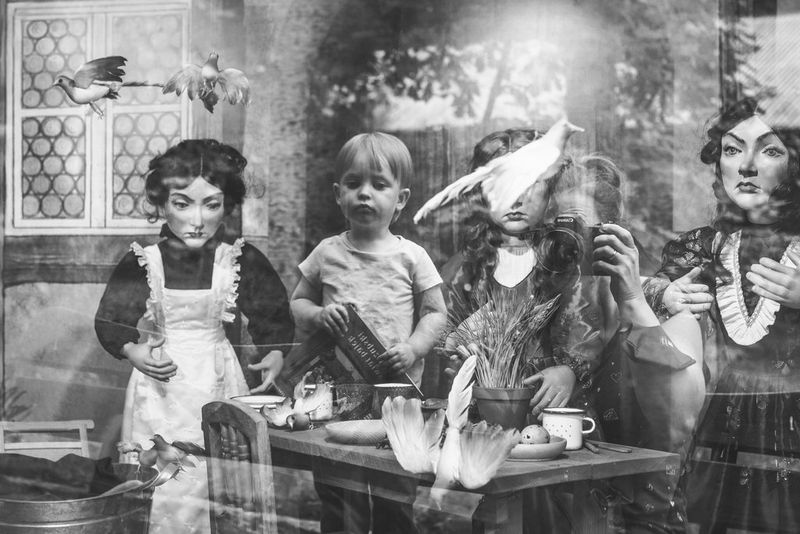 Črno-bel posnetek odseva deklice v oknu, ki daje vtis, da je deklica del vitrine z lutkami.