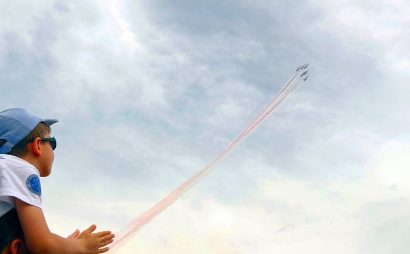Deček s čepico in sončnimi očali ploska med demonstracijskim preletom turških lovcev F-16, za katerimi se na nebu izriše rdeče-bela dimna sled.