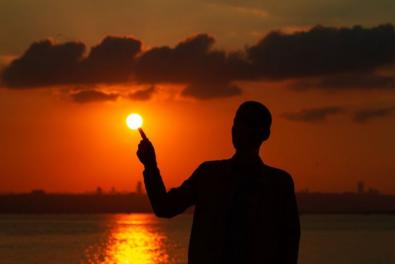 Silhueta moškega z iztegnjenim prstom, ki se navidezno dotika sonca.