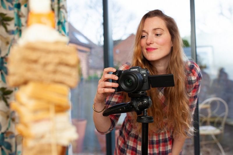 Een vrouw fotografeert een toren van eten met een camera op een statief bij een groot raam.