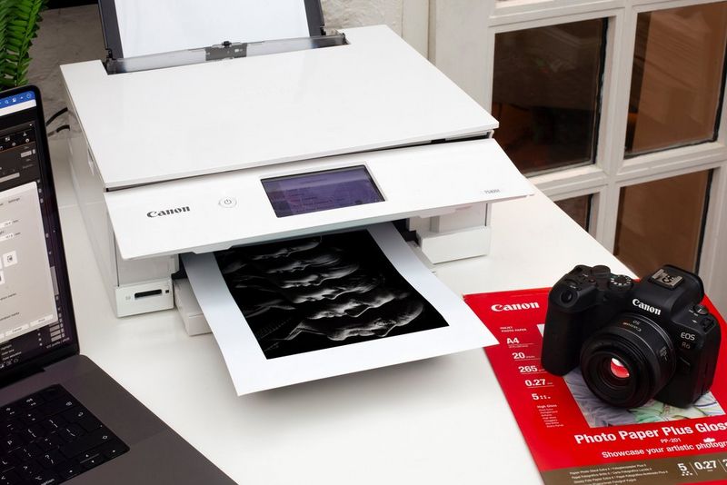 Tiskalnik družine Canon PIXMA TS8350 tiska črno-bel skupinski portret s Canonovim fotoaparatom in fotografskim papirjem na mizi ob njem.