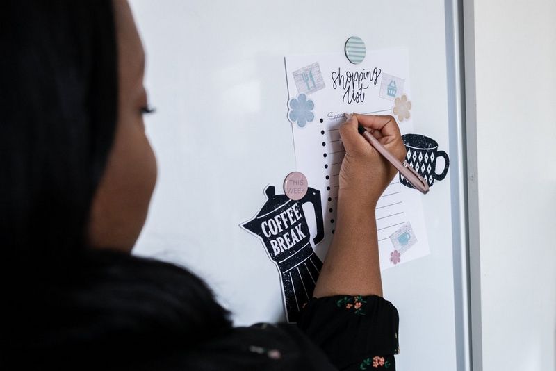 Женщина делает запись ручкой в красивом списке покупок, прикрепленном к дверце холодильника.