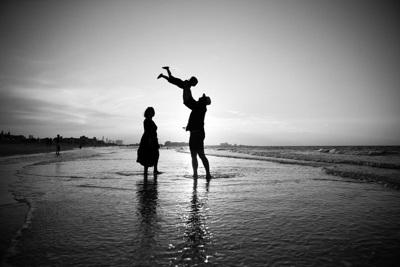 A black and white image of a family at the beach. The father is lifting his child into the air and all three are silhouetted against the sun.