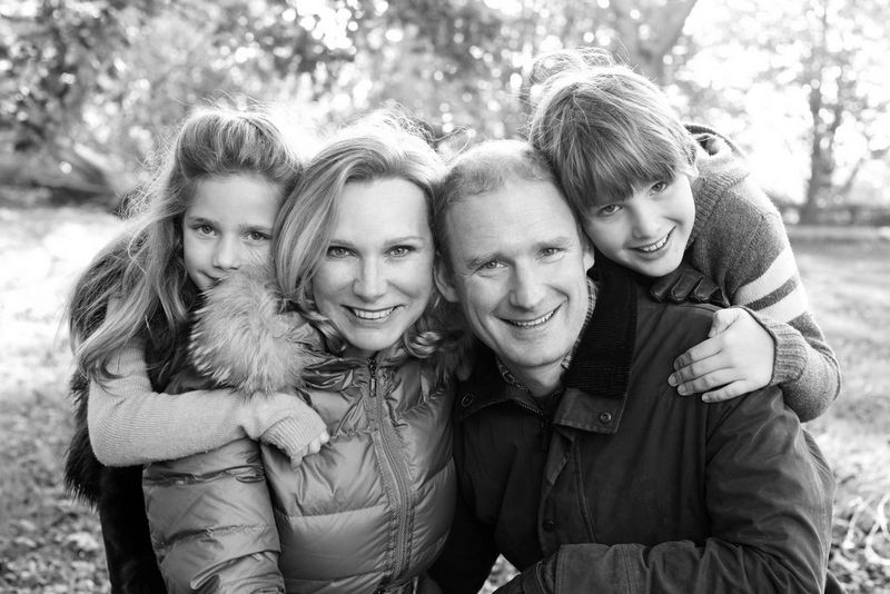 A black and white group portrait of a family posing outdoors, the parents crouched down and the two children leaning on their shoulders.