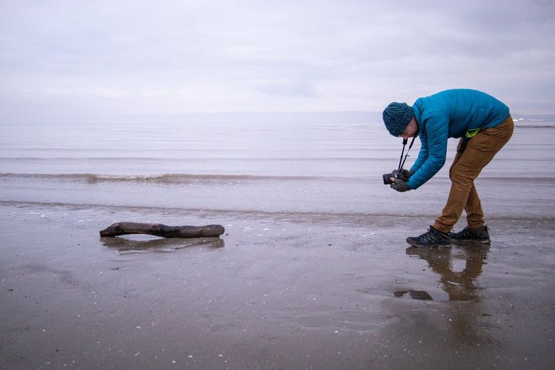A photographer bends down for a low-angle shot of a piece of driftwood on the shoreline.