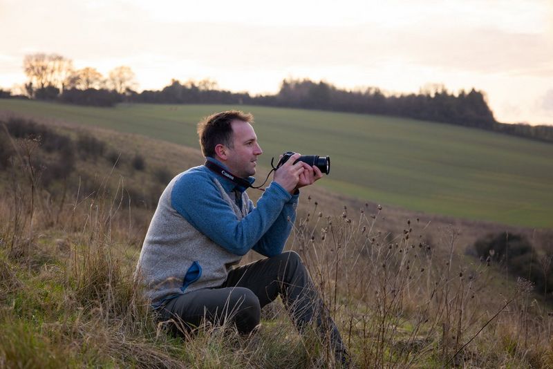 A landscape photographer looks through the viewfinder of a Canon EOS RP, steadying himself on his knee, to take a shot of rolling fields.