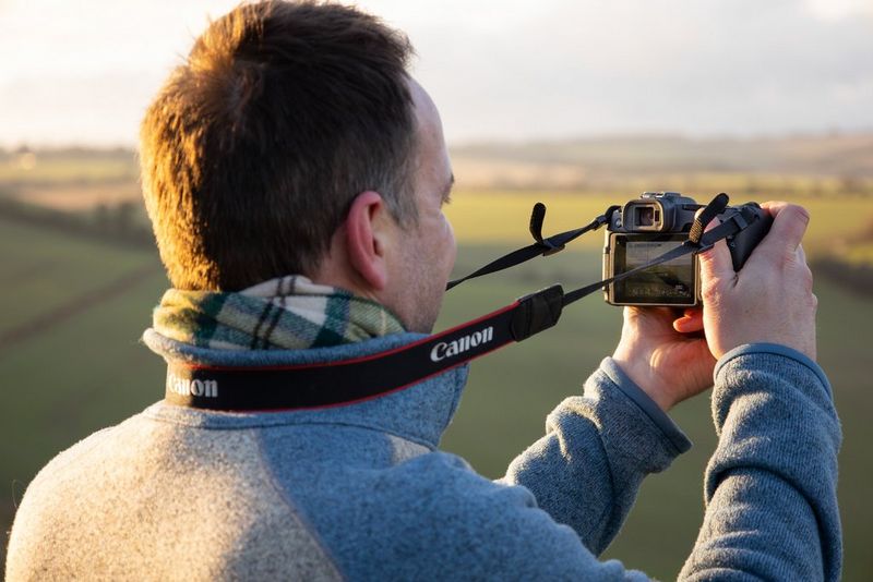 A landscape photographer holds a Canon EOS RP away from his body so the neck strap is taut.
