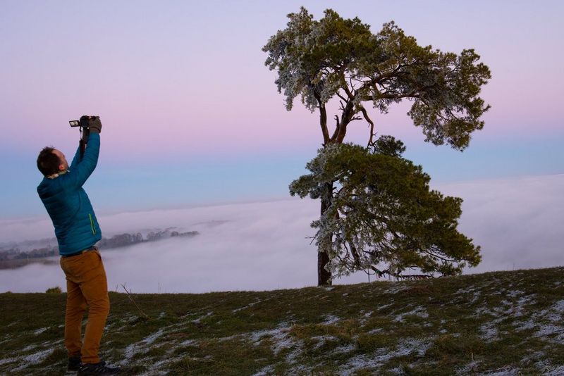 A landscape photographer holds a camera above his head to shoot a tree set against rolling clouds.