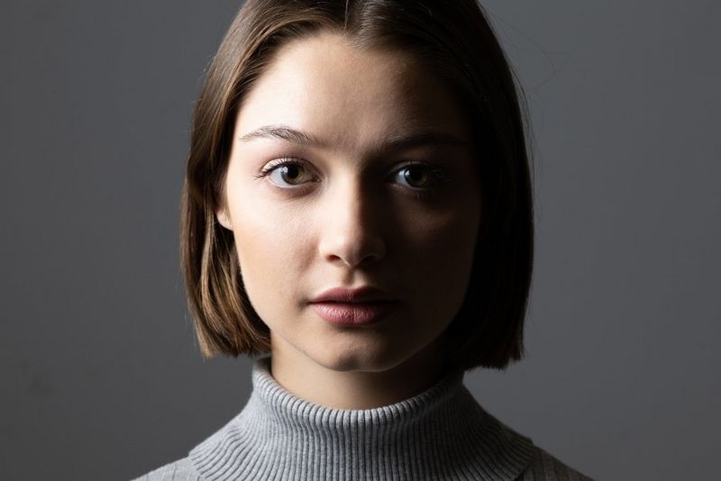 Portret van een vrouw in een grijze coltrui, tegen een grijze achtergrond, belicht van bovenaf.
