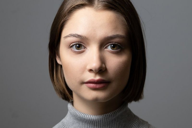 Portret van een vrouw in een grijze coltrui, tegen een grijze achtergrond met belichting vanaf linksboven, waardoor een lichtdriehoek op de rechterwang ontstaat.