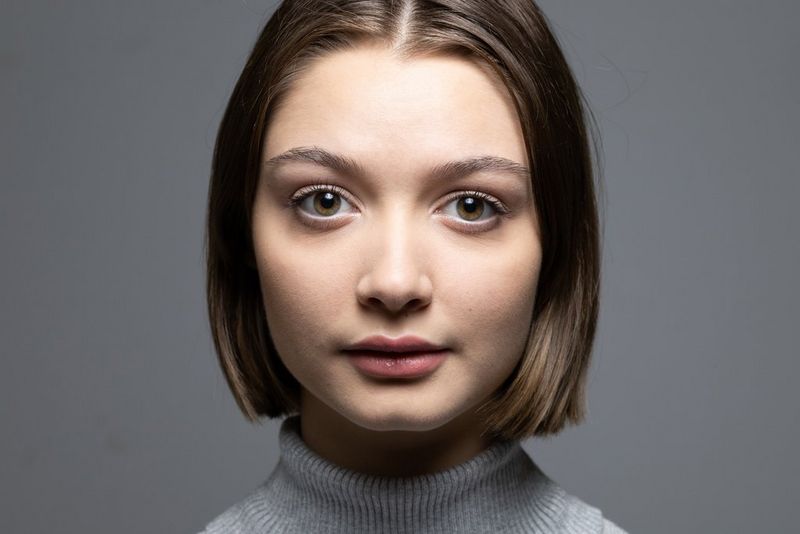 Portret van een vrouw in een grijze coltrui, tegen een grijze achtergrond, belicht van onderaf.