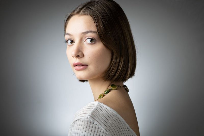 Portret van een vrouw met een grote groene ketting, tegen een grijze achtergrond en met een witte spotlight op de achtergrond.