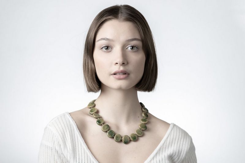 Portret van een vrouw met een grote, groene ketting en een witte top, tegen een witte achtergrond.