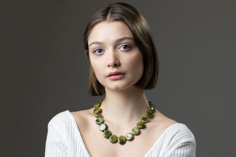 Portret van een vrouw met een grote groene ketting en een witte top, tegen een donkergrijze achtergrond.