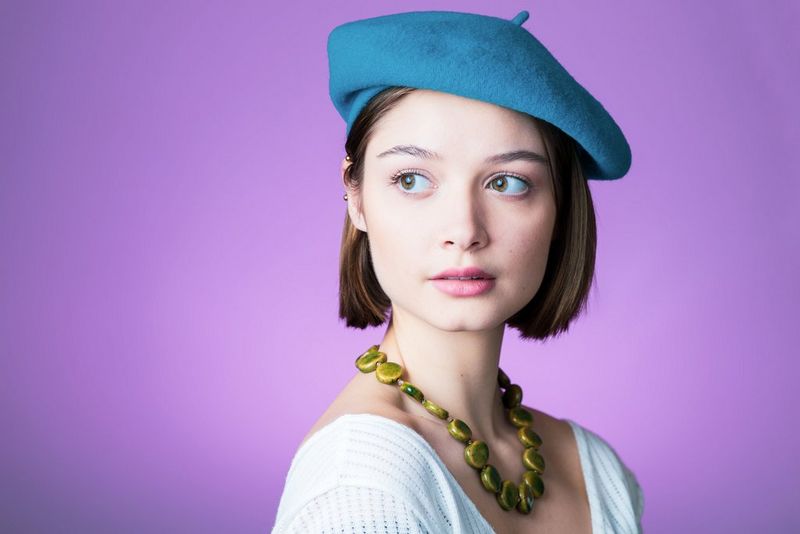 Portret van een vrouw met een blauwe baret, tegen een paarse achtergrond.
