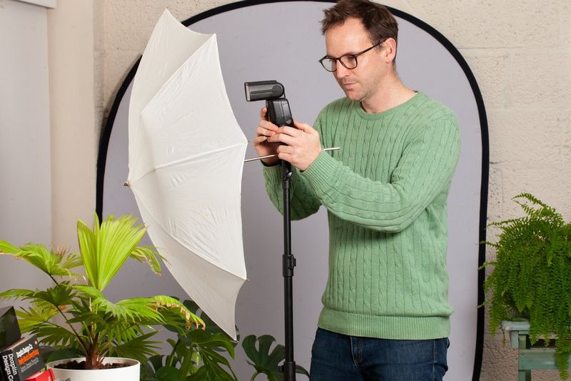 Een man in een groene trui, met een Canon-flitser op een statief dat naar een witte paraplu is gericht.