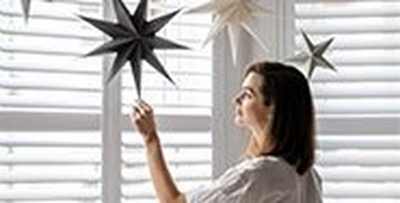 A woman standing in front of shuttered windows touches a paper star hanging from the ceiling.