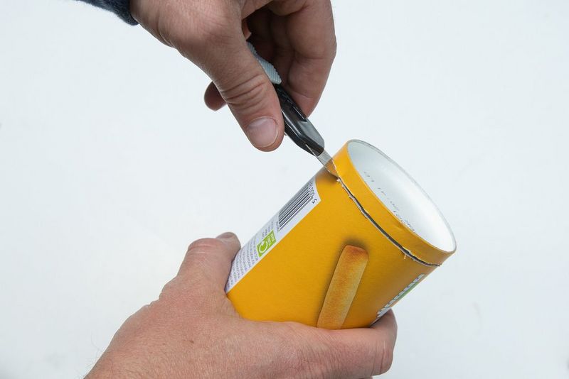 Мужчина держит в руке тубус для закусок и отрезает от него дно с помощью канцелярского ножа.