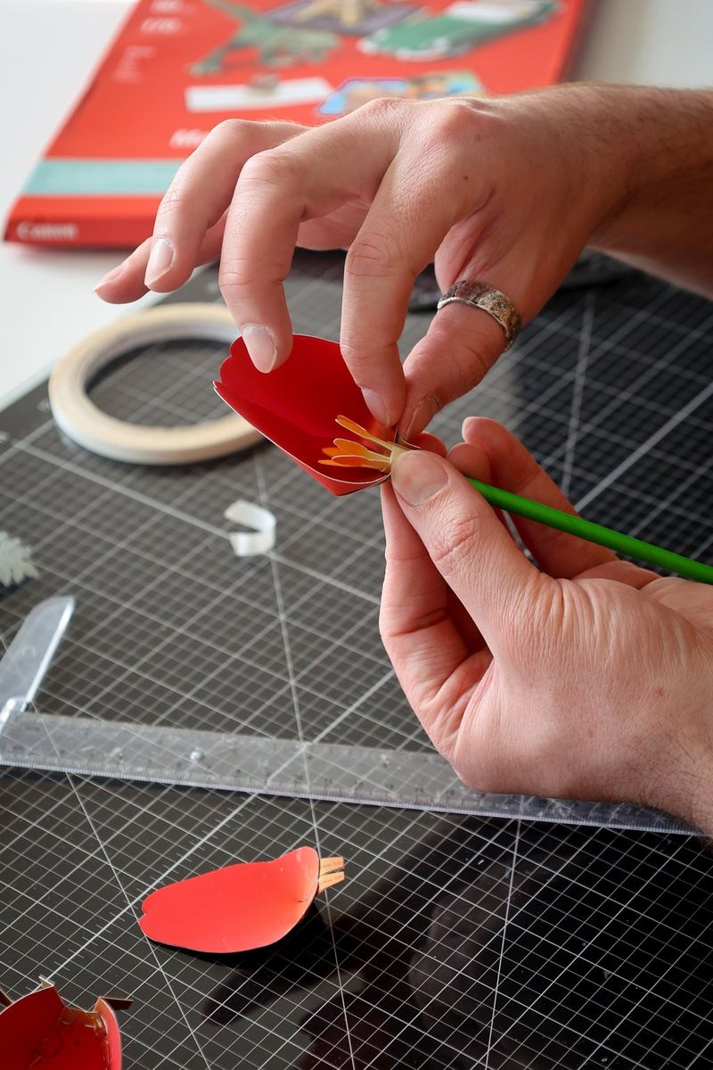 A pair of hands places the first papercraft petal on to the stem of a flower. Further petals can be seen on the cutting board below.