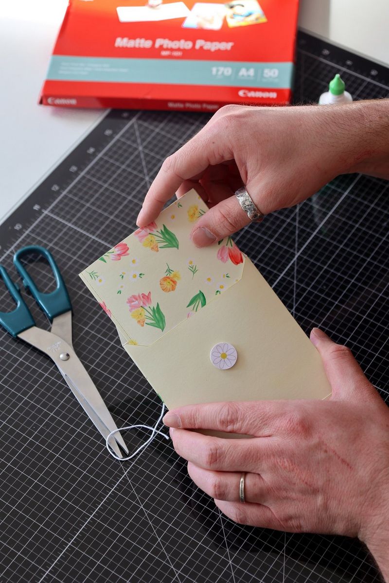 A pair of hands places a card inside a handmade envelope. On the table below are scissors, glue and a pack of Canon paper.