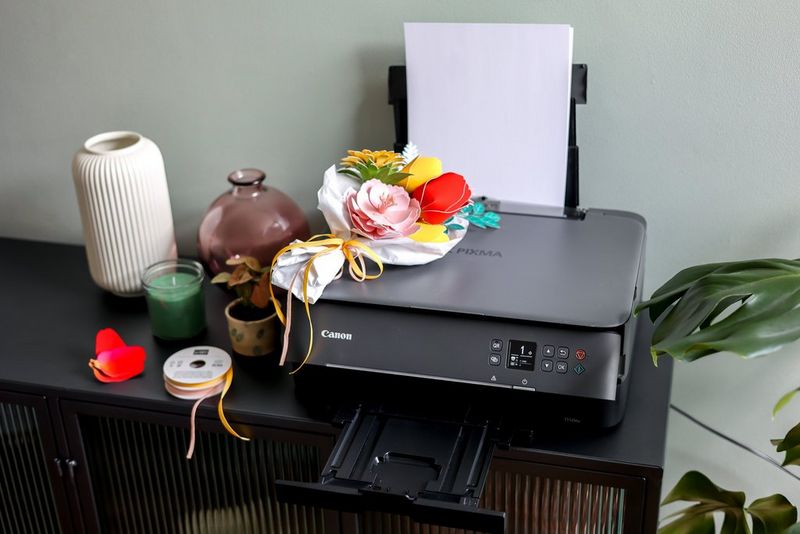 A Canon PIXMA printer and several jars and candles are positioned on a black desk. On top of the printer is a handcrafted bouquet of paper flowers. 