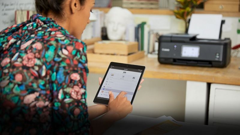 A woman with her back to the camera uses a tablet. A Canon printer is on a desk in the background.