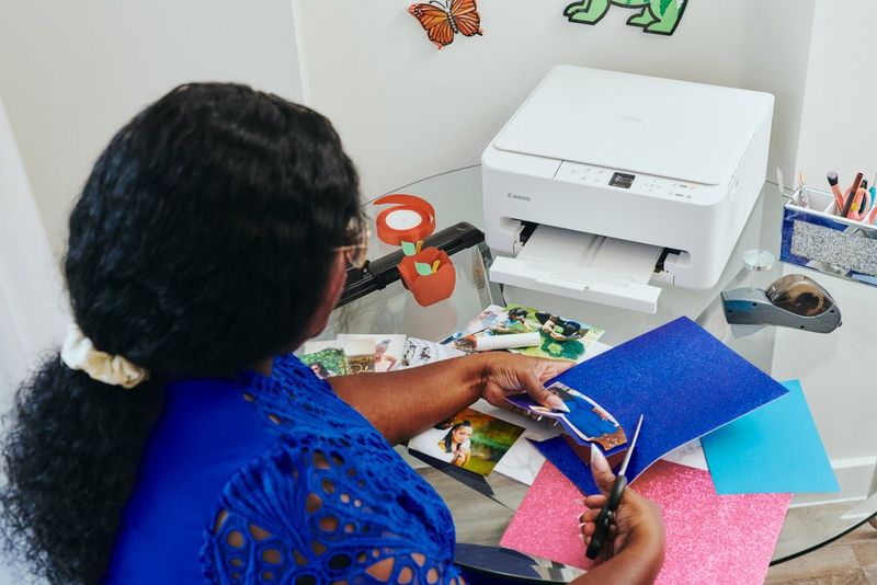 Kunsthåndværkeren Juliet Amma klipper et fotoprint og farvet papir ud, med flere fotoprints og en Canon PIXMA-printer på bordet foran sig.