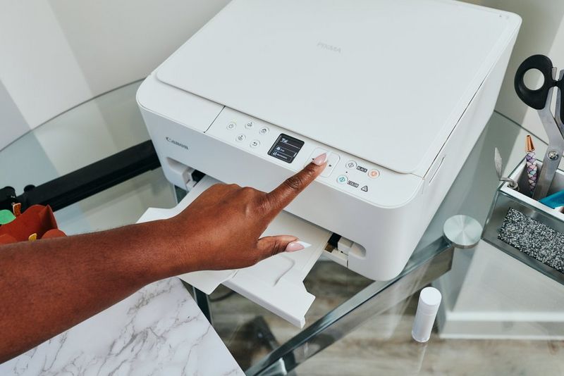 Een hand drukt op een knop van een Canon PIXMA-printer die op een glazen tafel staat.