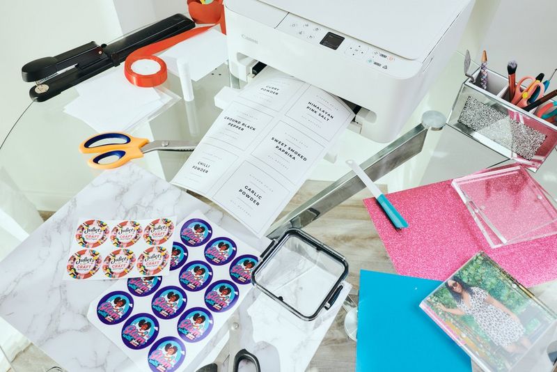 Geprinte stickeretiketten, kleurrijke kaartjes en andere geprinte items liggen op een glazen tafel met verschillende knutseltools en een Canon PIXMA-printer.