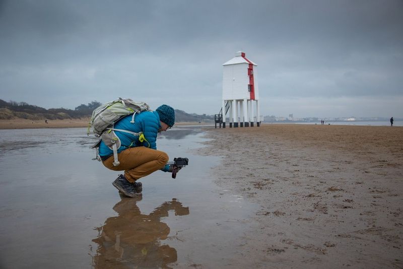 A photographer crouching on a beach to take a low angle shot of a Victorian lighthouse.