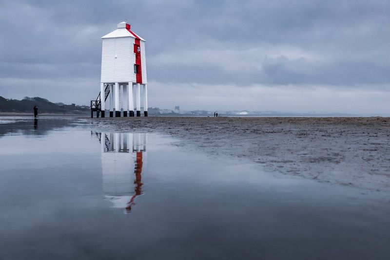 A Victorian lighthouse is reflected in the water on the sand.