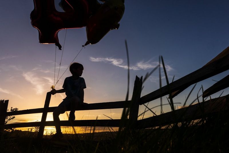 Un bambino seduto su una recinzione che tiene un mazzo di palloncini di fronte a un tramonto. Scatto realizzato con Canon EOS R6 e obiettivo Canon RF 24-105mm F4-7.1 IS STM.