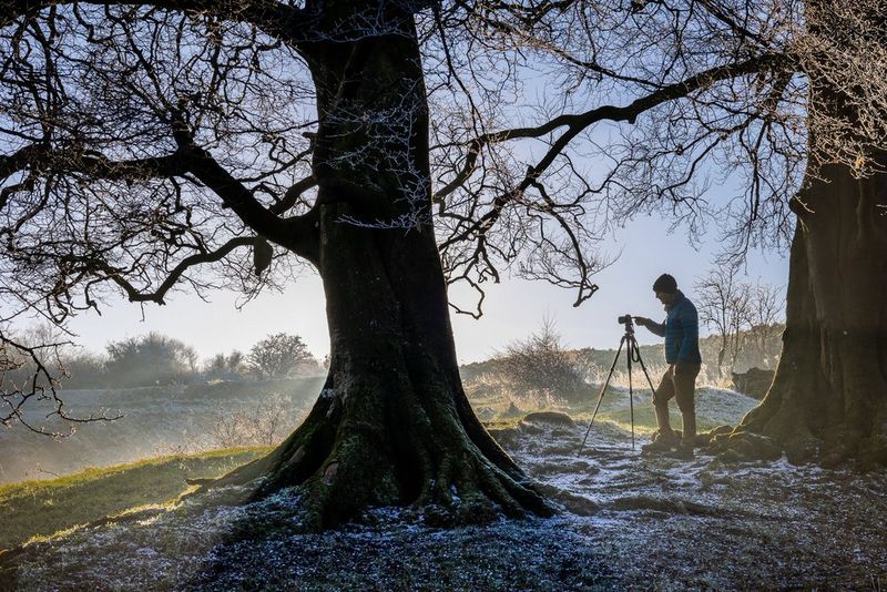 Un uomo si trova in una foresta in una giornata invernale, con la sua fotocamera Canon posizionata su un treppiede.