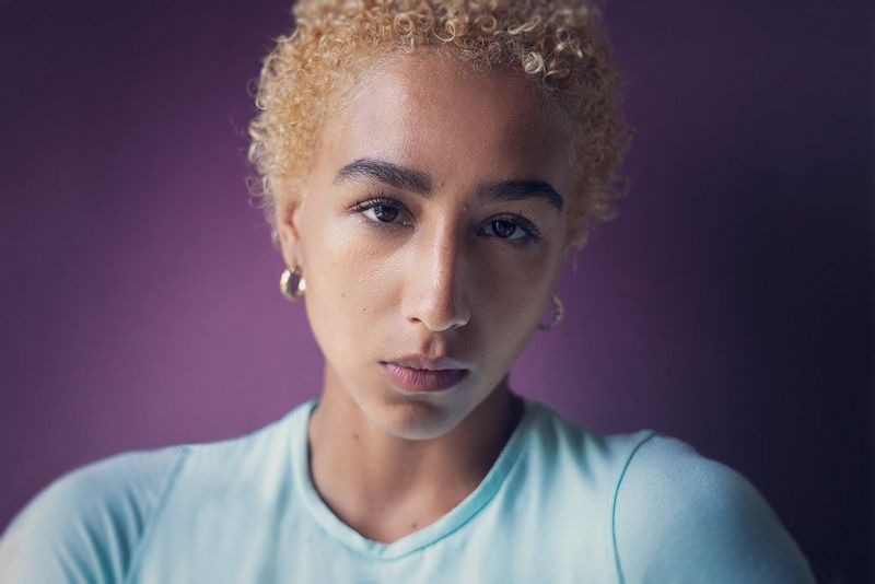 Un ritratto, illuminato lateralmente e realizzato con Canon EOS R6 con un obiettivo Canon RF 50mm F1.8 STM, di una persona con capelli biondi e ricci che guarda direttamente verso la fotocamera.